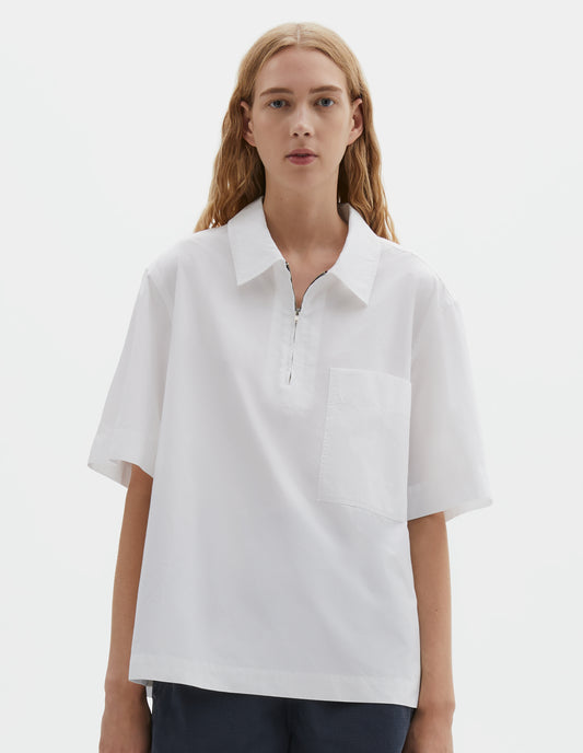 Cropped body image, front view of model wearing relaxed short-sleeved white shirt with wide stitch-through placket with half zip, patch chest pocket and side split hem. Styled with dark blue shorts.