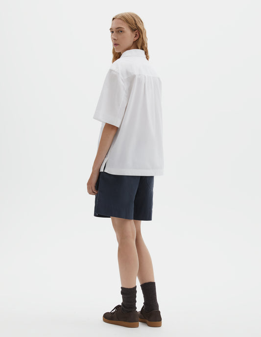 Full body image, back view of model wearing relaxed short-sleeved white shirt with side split hem and wide box pleat on back yoke. Styled with dark blue shorts, dark brown socks and dark brown nubuck trainers.