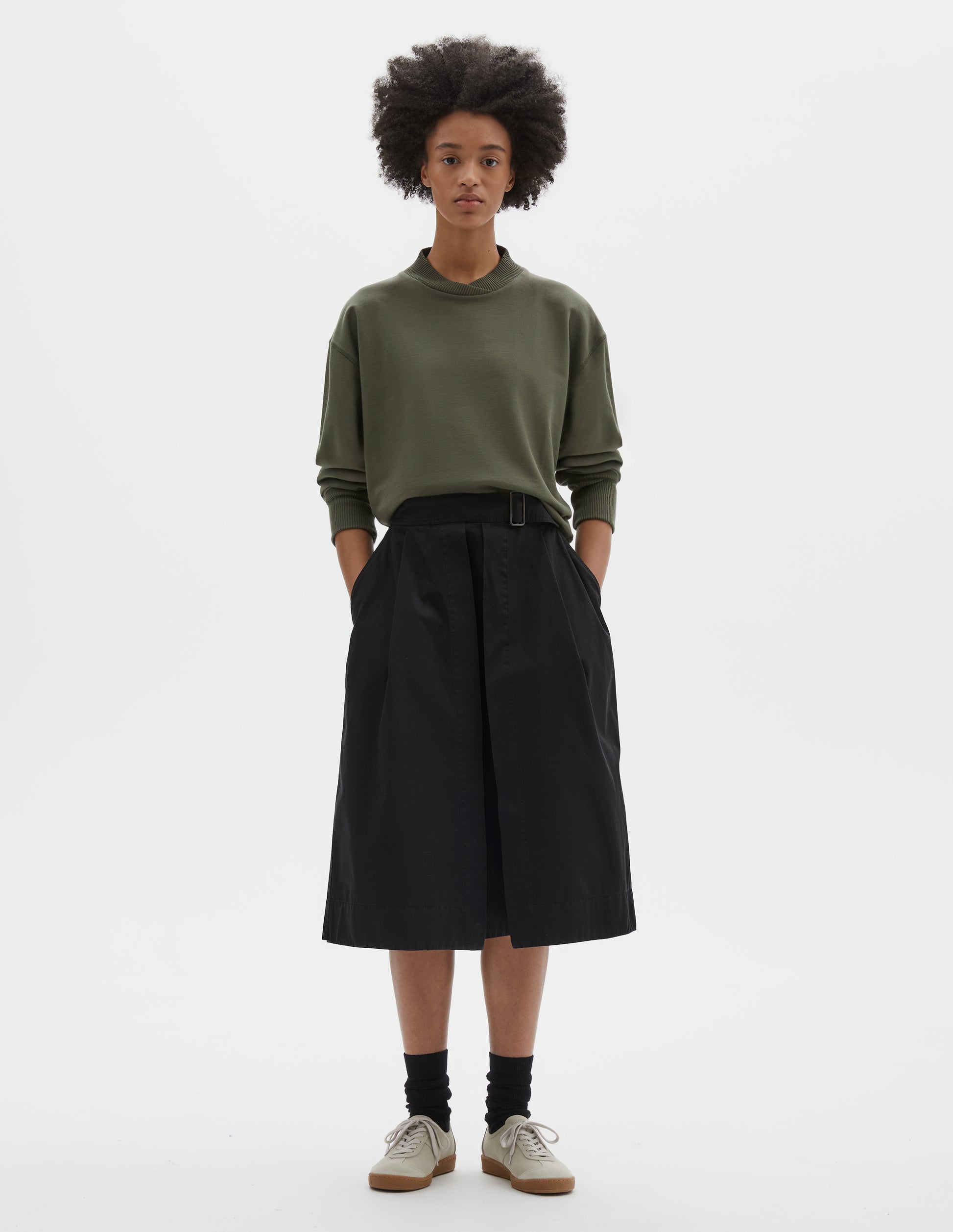 Full body image, front view of model wearing black cotton twill skirt, with wide crossover waistband, button fastening slide adjuster and slant pockets. Styled with green cotton sweatshirt, black socks and beige suede trainers.
