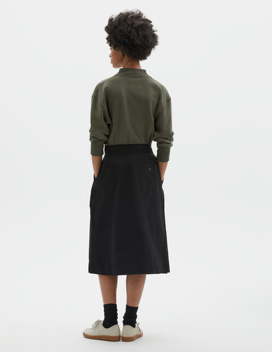 Full body image, back view of model wearing black cotton twill skirt, with wide crossover waistband and stitch through welt back pocket. Styled with green cotton sweatshirt, black socks and beige suede trainers.