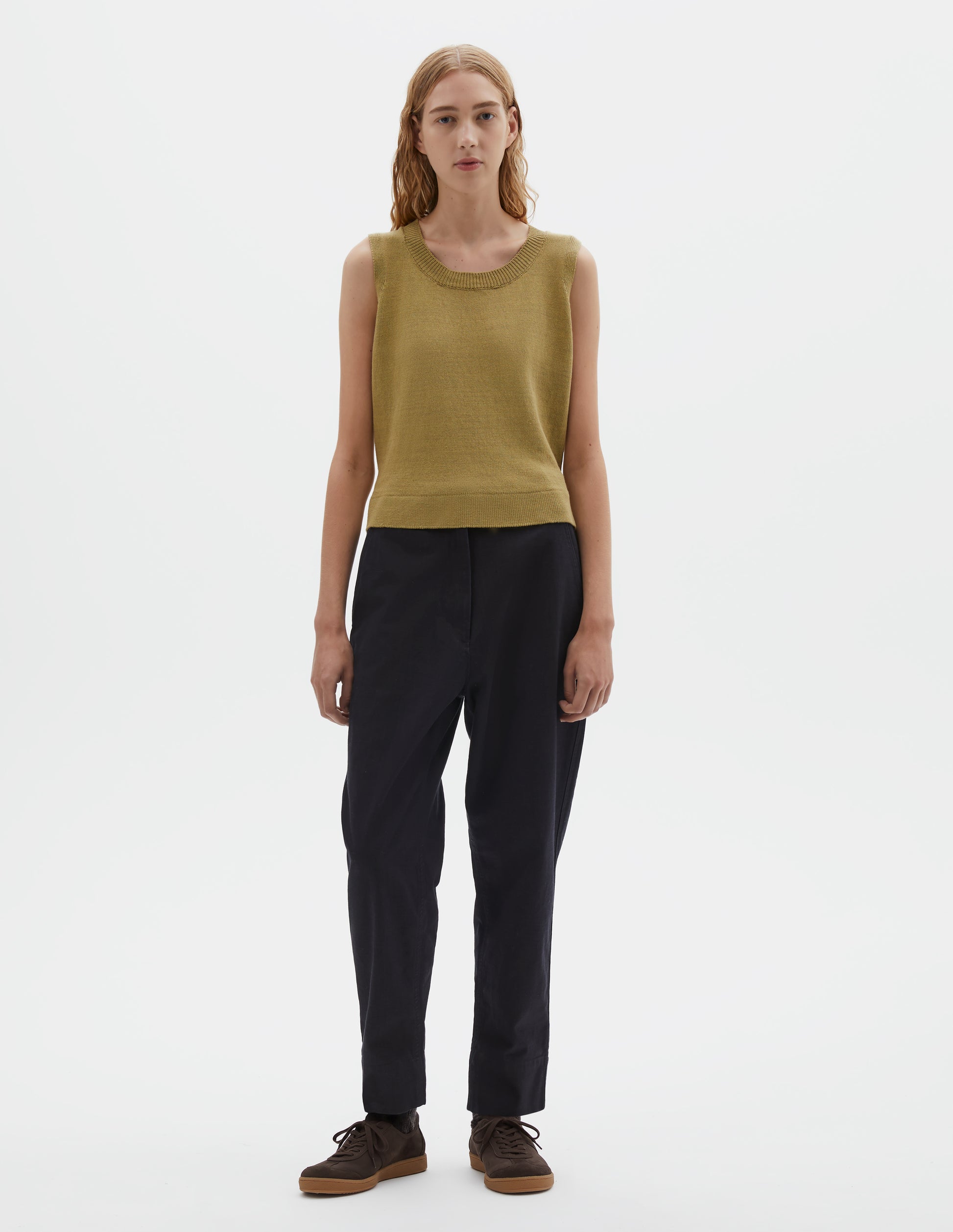 Full body image, front view of model wearing dark blue trousers with zip fly and corozo button fastening, slant side pockets with ticket pocket detail and deep stitch hem. Styled with yellow knitted tank top and brown suede trainers.