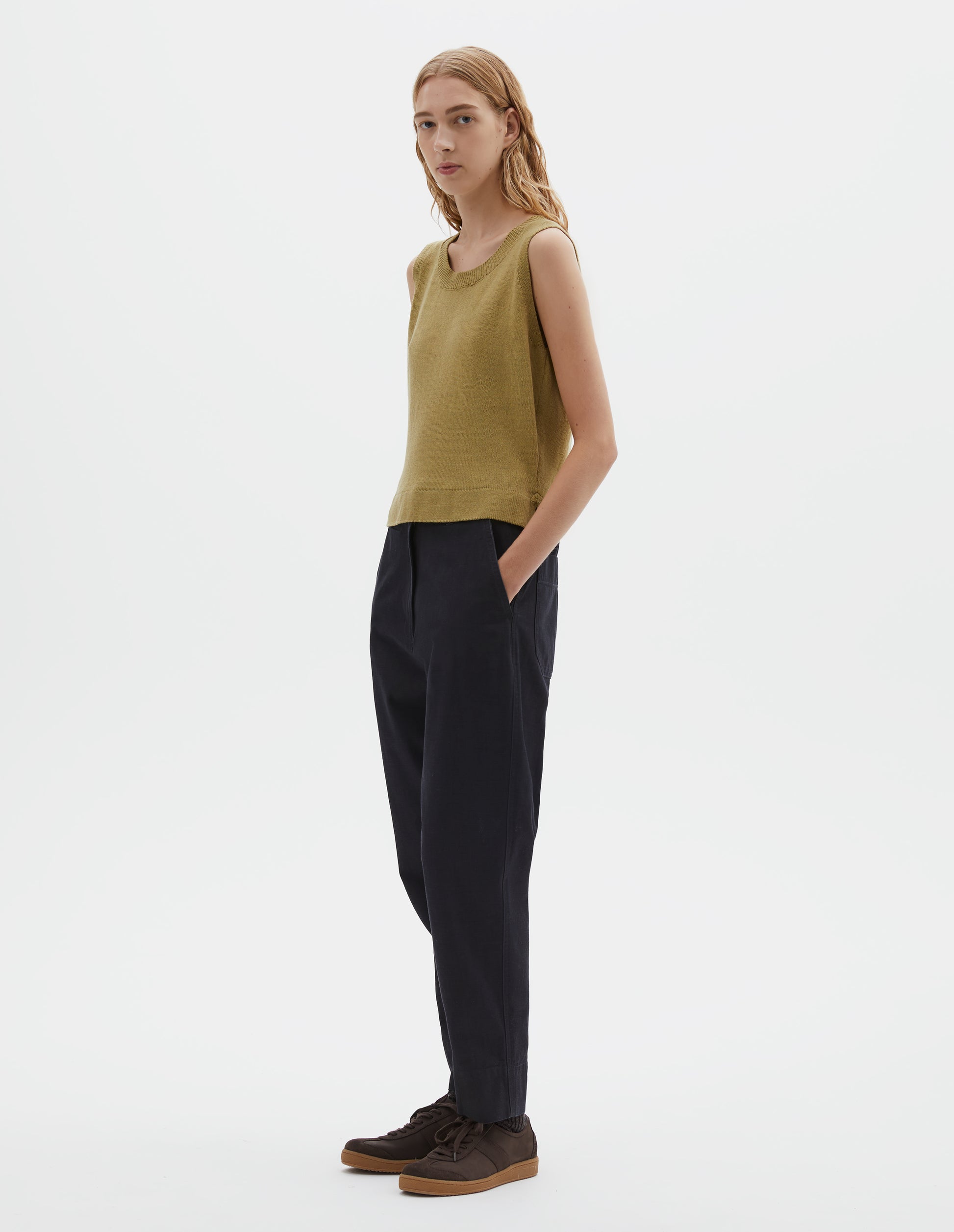 Full body image, front view of model wearing regular fit yellowish green knitted tank top with single rib crew neck and side split rib hem. Styled with dark blue trousers and dark brown nubuck trainers.