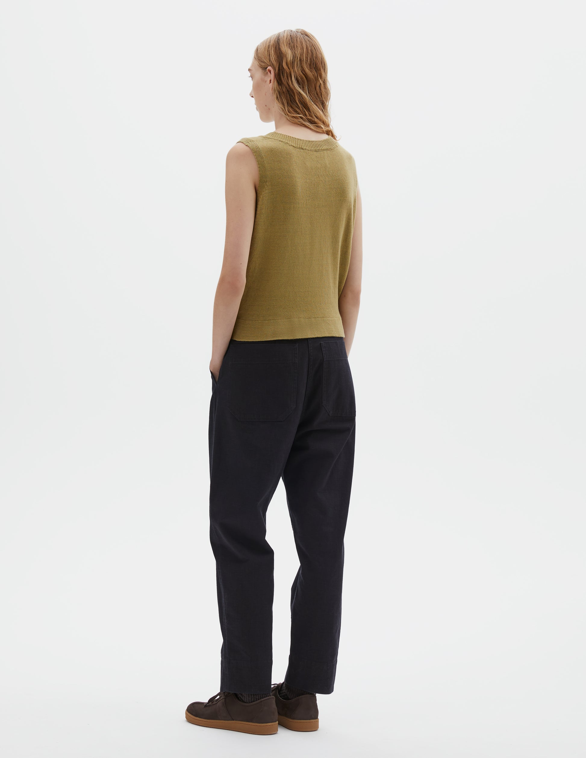 Full body image, back view of model wearing dark blue trousers with belt loops, corozo button tab side adjusters and patch back pockets. Styled with yellow knitted tank top and brown suede trainers.