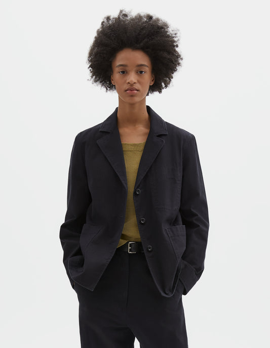 Cropped body image, front view of model wearing indigo twill single breasted blazer, with two front patch pockets, fold back split cuff, curved hem and corozo button closure. Styled over green vest, with matching indigo twill trousers and black leather belt.