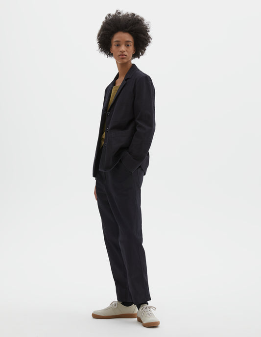 Full body image, front view of model wearing indigo twill single breasted blazer, with two front patch pockets, fold back split cuff, curved hem and corozo button closure. Styled over green vest, with matching indigo twill trousers, black socks and beige suede trainers.
