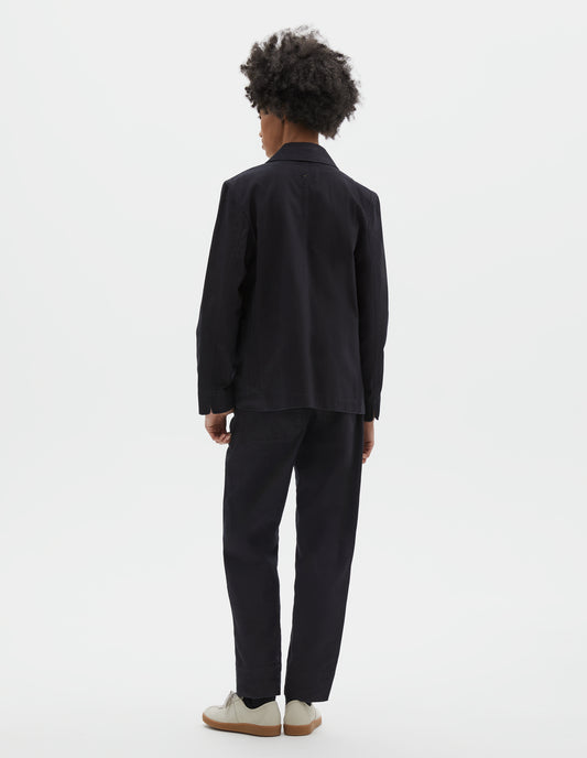 Full body image, back view of model wearing indigo twill single breasted blazer, with fold back split cuff and curved hem. Styled with matching indigo twill trousers, black socks and beige suede trainers.