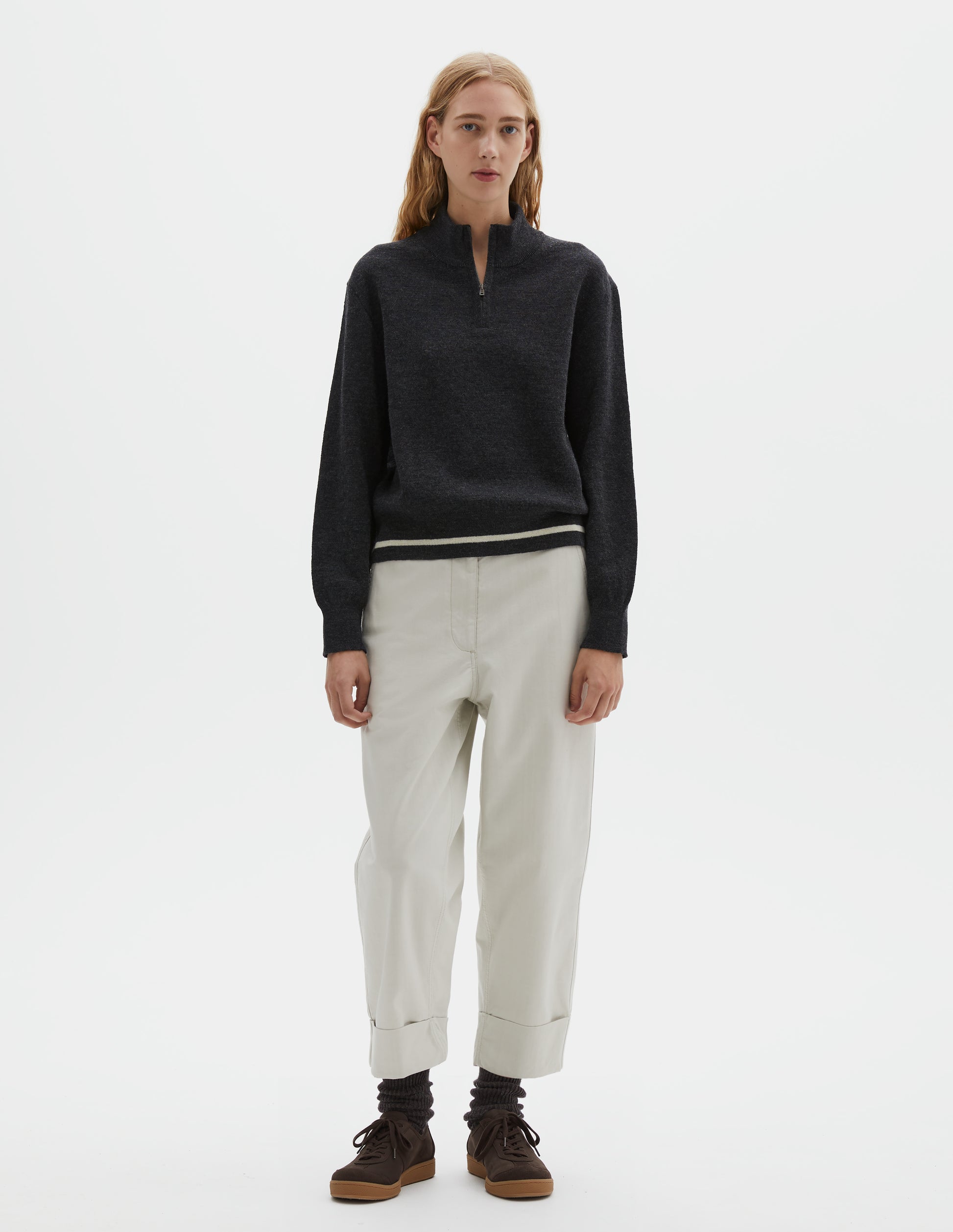 Full body image, front view of model wearing beige cotton drill trousers with slant pockets, corozo button detailing on waistband and turn up hems. Styled with half zip jumper, dark brown socks with dark brown nubuck trainers.
