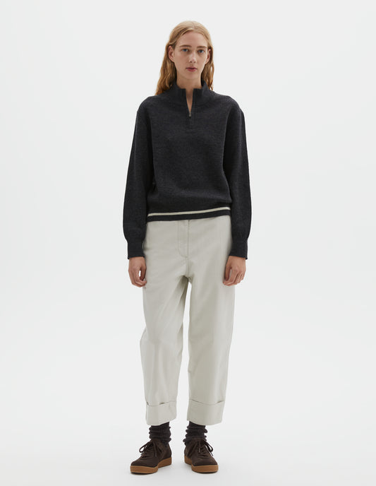 Full body image, front view of model wearing beige cotton drill trousers with slant pockets, corozo button detailing on waistband and turn up hems. Styled with half zip jumper, dark brown socks with dark brown nubuck trainers.