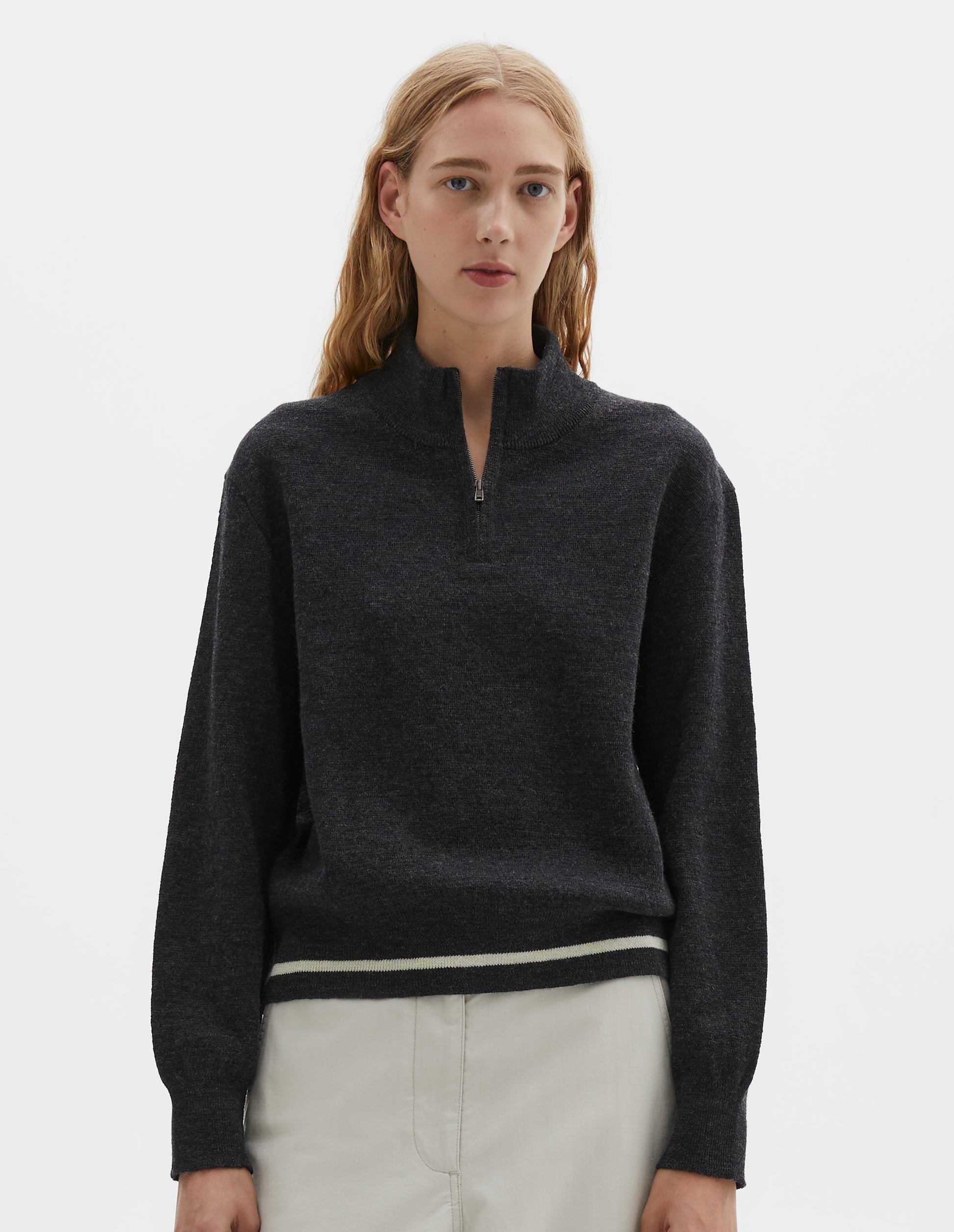 Cropped body image, front view of model wearing dark grey wool cotton half zip sweater with contrast rib on neck and cuffs, and contrast white line around ribbed hem. Styled with light grey trousers.