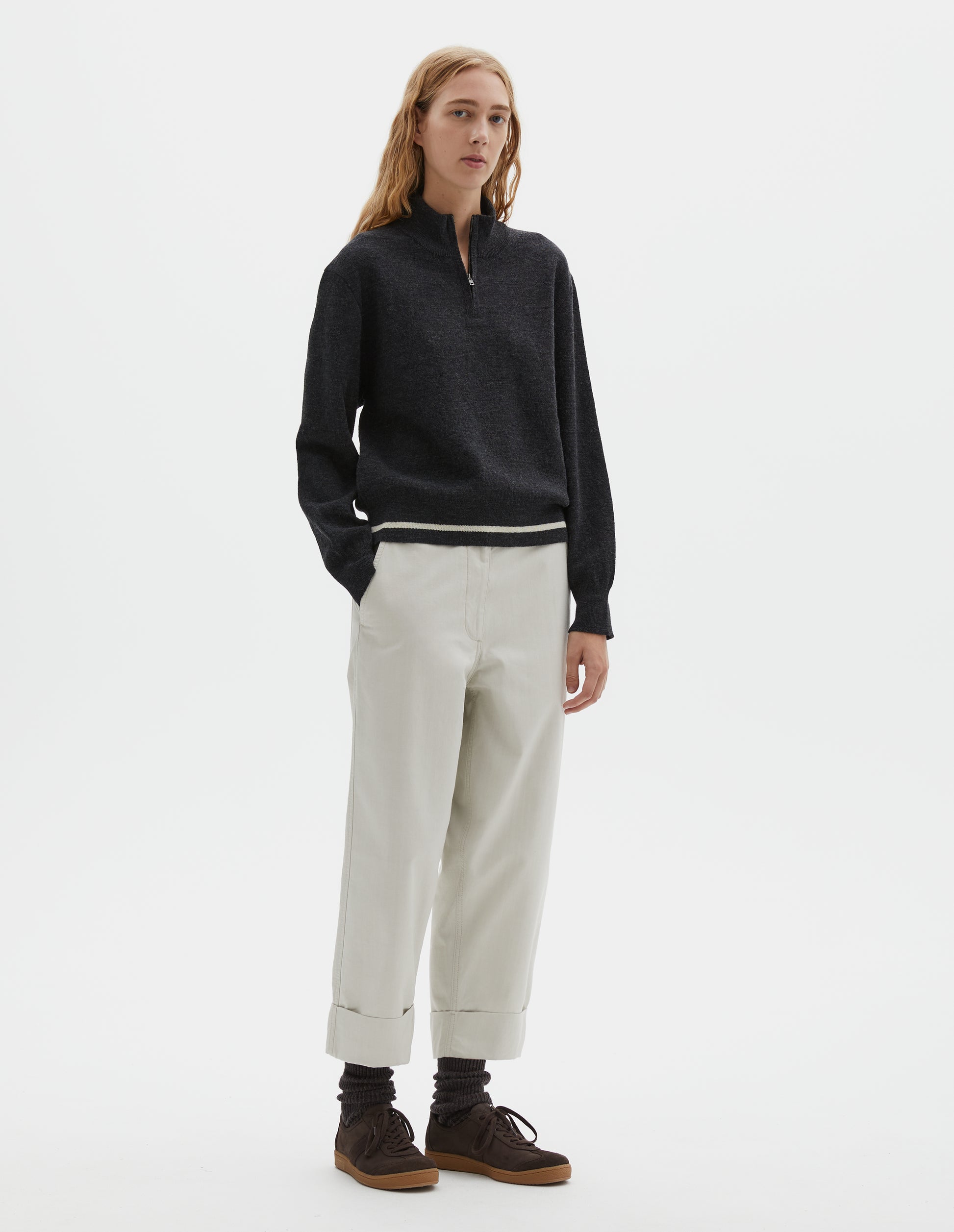 Full body image, front view of model wearing dark grey wool cotton half zip sweater with contrast rib on neck and cuffs, and contrast white line around ribbed hem. Styled with light grey cropped trousers and dark brown socks with dark brown nubuck trainers.