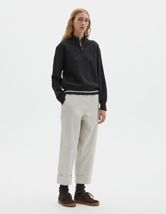 Full body image, front view of model wearing dark grey wool cotton half zip sweater with contrast rib on neck and cuffs, and contrast white line around ribbed hem. Styled with light grey cropped trousers and dark brown socks with dark brown nubuck trainers.