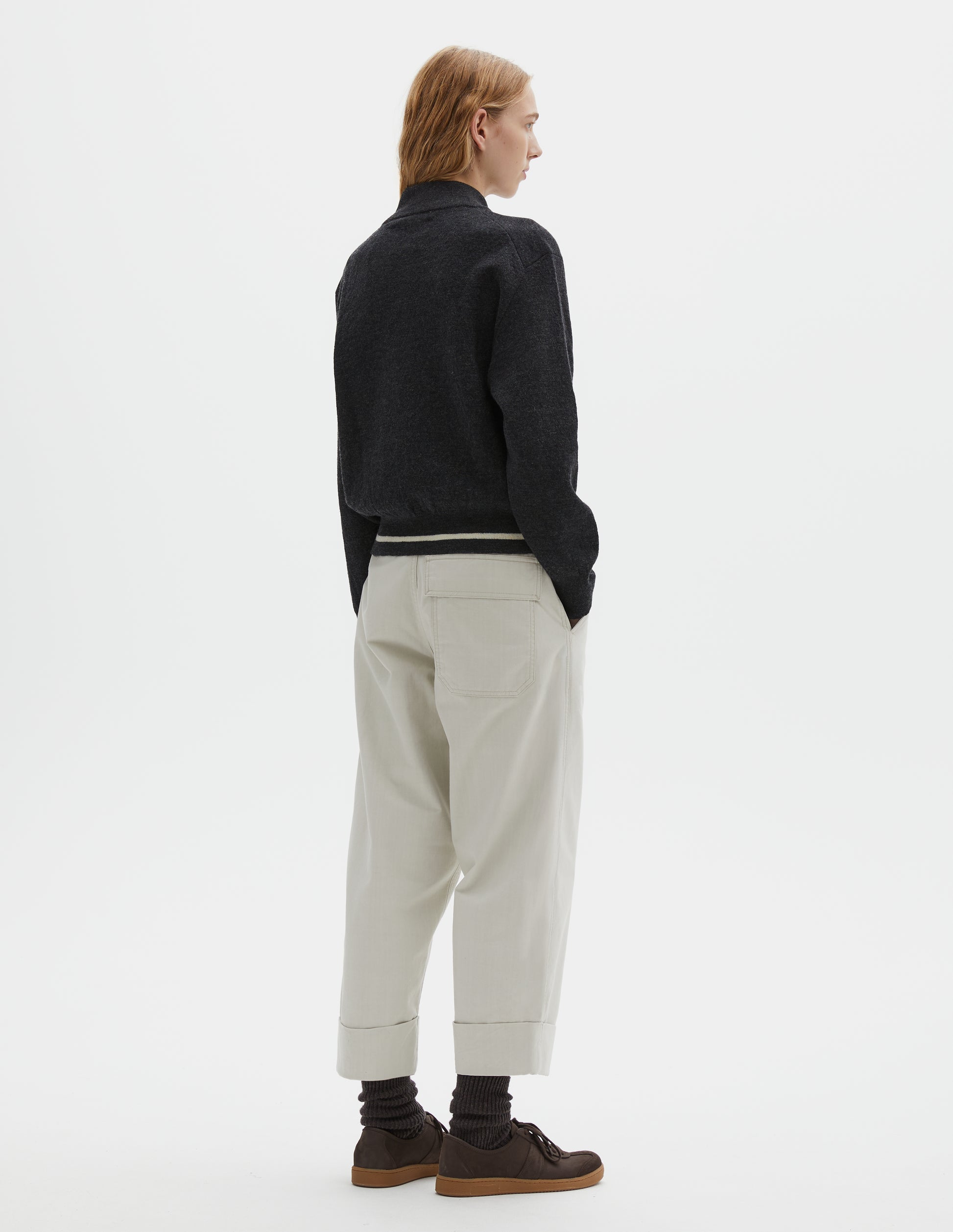 Full body image, back view of model wearing dark grey wool cotton sweater with contrast rib on neck and cuffs, and contrast white line around ribbed hem. Styled with light grey cropped trousers and dark brown socks with dark brown nubuck trainers.