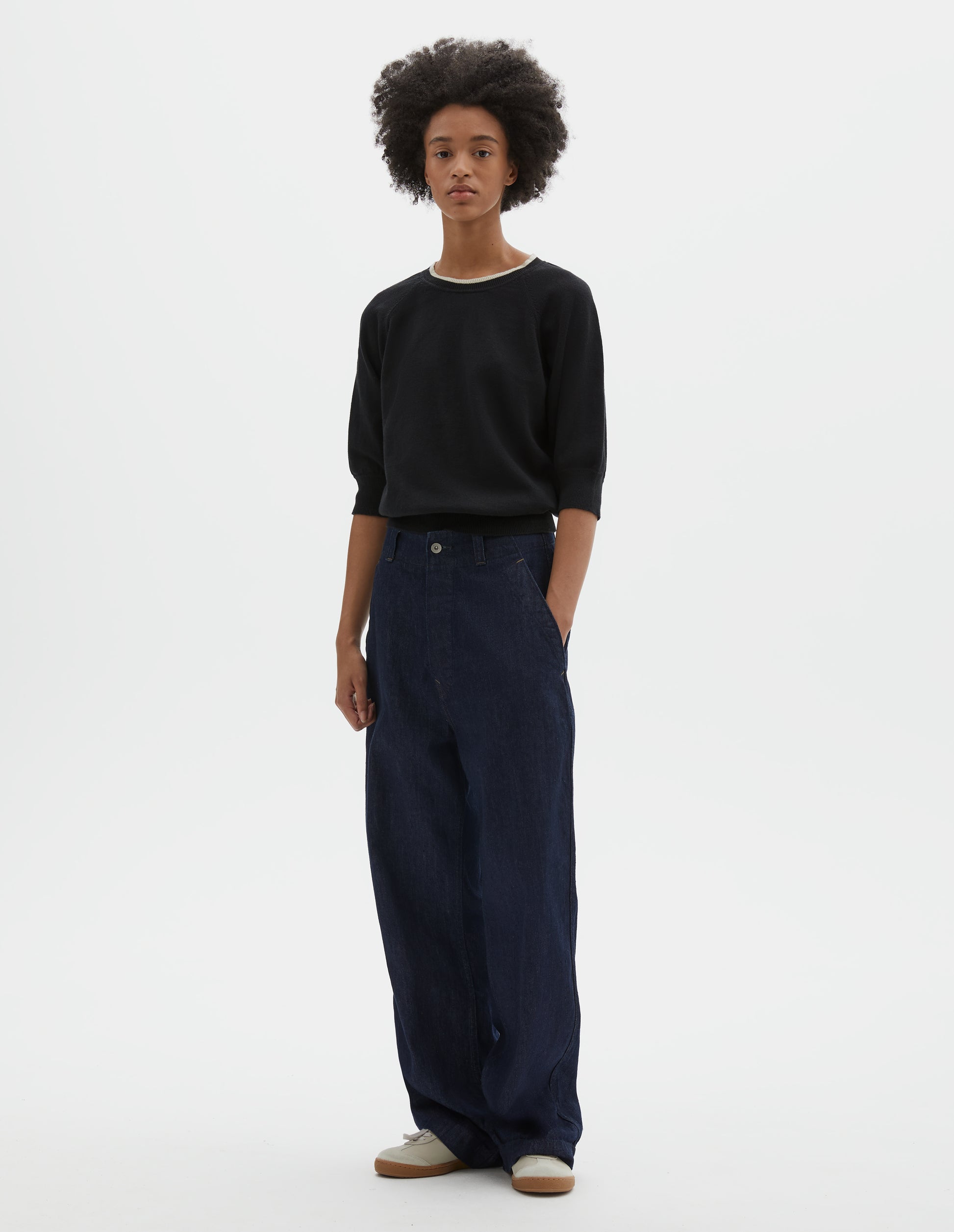 Full body image, front view of model wearing black short sleeve knitted t-shirt, with contrast white stitching on neckline, raglan sleeve, flatlock stitch seam detailing and contrast rib on sleeve and hem. Styled with dark navy blue denim jeans and white suede shoes with gum sole.