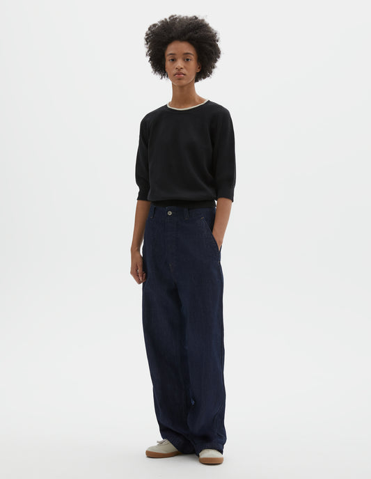 Full body image, front view of model wearing black short sleeve knitted t-shirt, with contrast white stitching on neckline, raglan sleeve, flatlock stitch seam detailing and contrast rib on sleeve and hem. Styled with dark navy blue denim jeans and white suede shoes with gum sole.