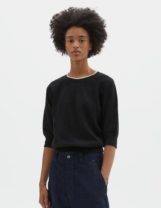 Cropped body image front view of model wearing black short sleeve knitted t-shirt, with contrast white stitching on neckline, raglan sleeve, flatlock stitch seam detailing and contrast rib on sleeve and hem. Styled with dark navy blue denim jeans.