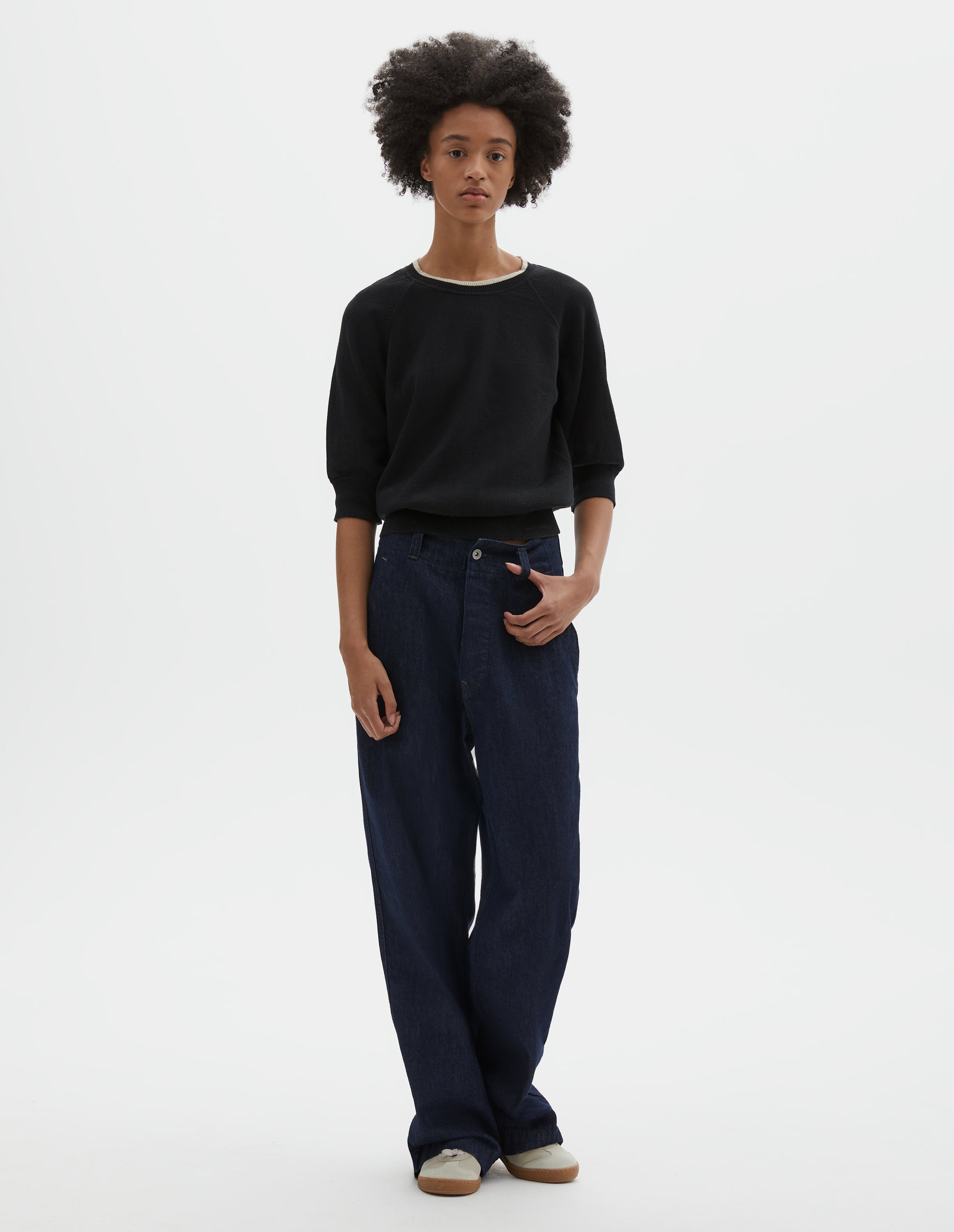 Full body image, front view of model wearing indigo blue wide leg light denim twill jeans. With metal button closure, button through waistband and slant side pockets. Styled with black short sleeve knitted top and beige suede trainers.