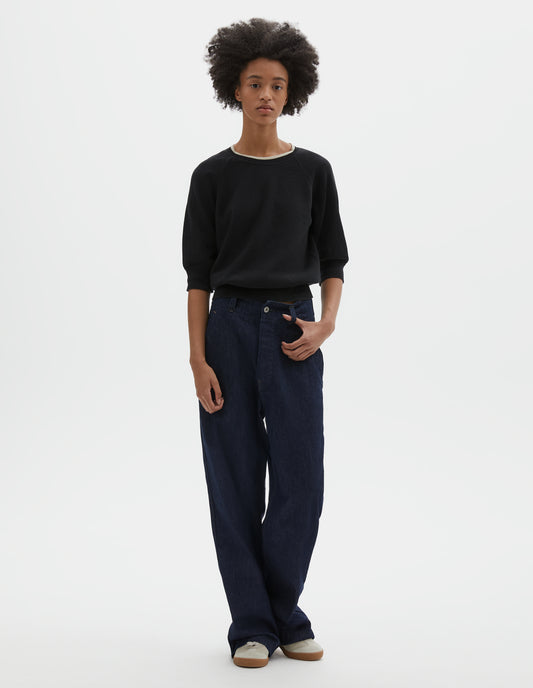 Full body image, front view of model wearing indigo blue wide leg light denim twill jeans. With metal button closure, button through waistband and slant side pockets. Styled with black short sleeve knitted top and beige suede trainers.