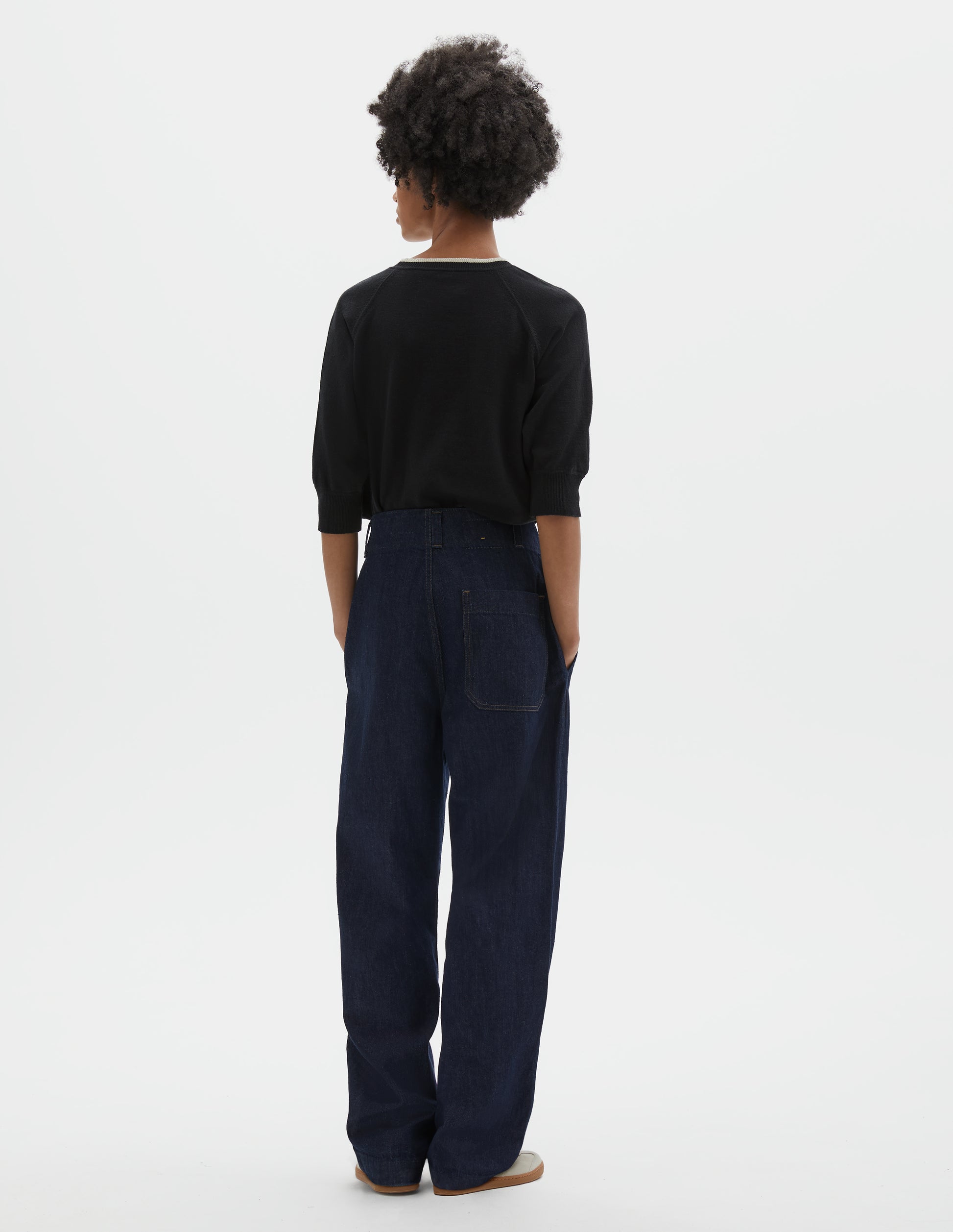 Full body image, back view of model wearing indigo blue wide leg light denim twill jeans. With slant side pockets and patch back pocket. Styled with black short sleeve knitted top and beige suede trainers.