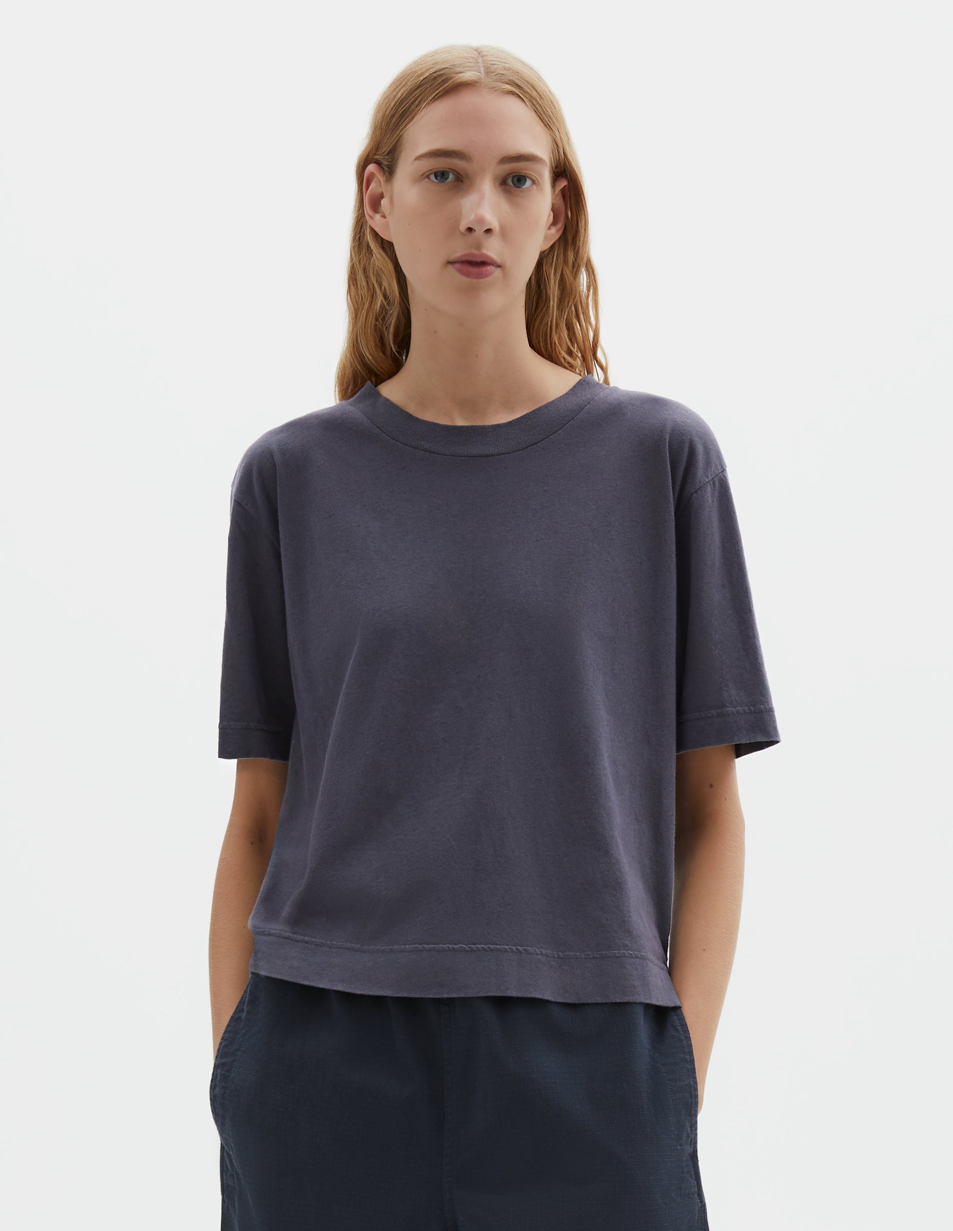Cropped body image, side view of model wearing greyish blue cotton and linen t-shirt, with vent split hem. Styled with navy blue shorts.