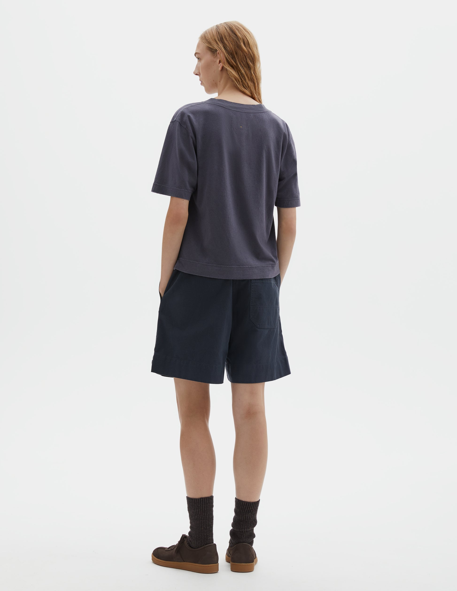 Full body image, back view of model wearing greyish blue cotton and linen t-shirt, with vent split hem. Styled with navy blue shorts, black socks and brown suede trainers.