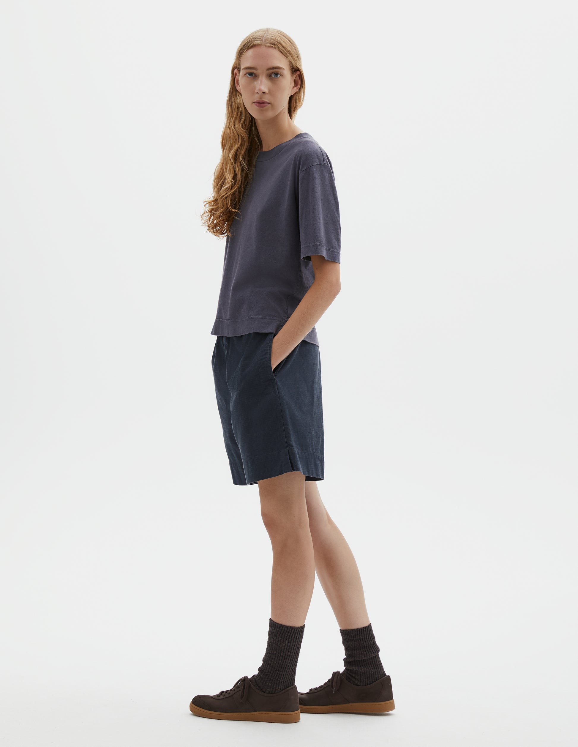 Full body image, side view of model wearing greyish blue cotton and linen t-shirt, with vent split hem. Styled with navy blue shorts, black socks and brown suede trainers.