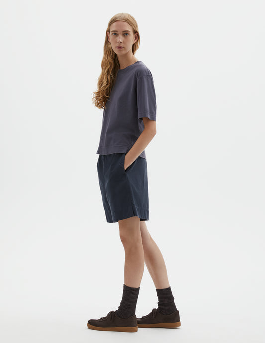 Full body image, side view of model wearing greyish blue cotton and linen t-shirt, with vent split hem. Styled with navy blue shorts, black socks and brown suede trainers.