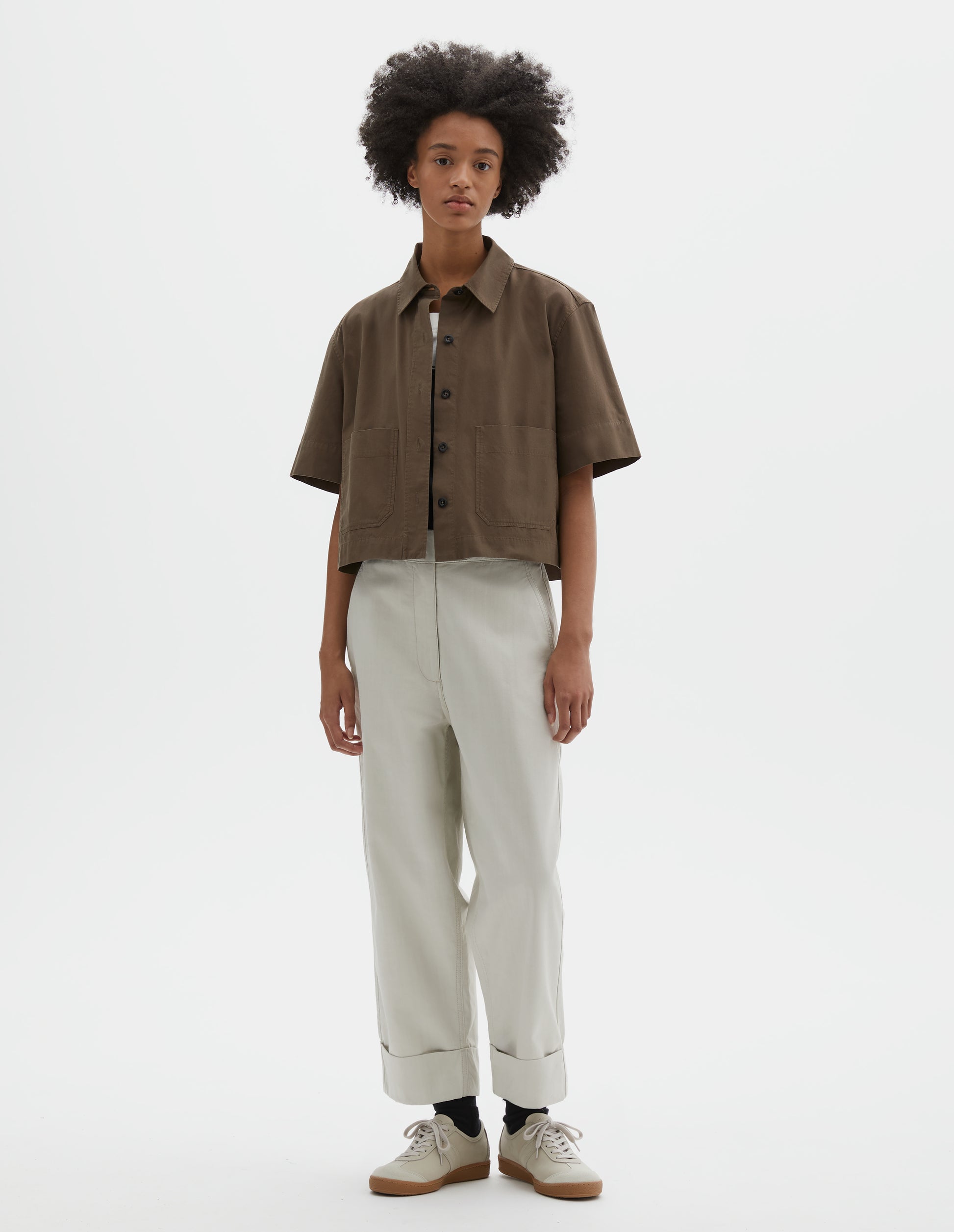 Full body image, front view of model wearing greenish brown cotton short sleeve shirt, with raised placket, patch pockets, stepped side hem and corozo brace buttons. Styled over white t-shirt and white trousers, black socks and beige nubuck trainers.