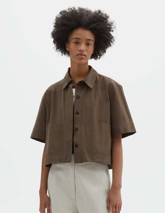 Cropped body image, front view of model wearing greenish brown cotton short sleeve shirt, with raised placket, patch pockets, stepped side hem and corozo brace buttons. Styled over white t-shirt and white trousers.