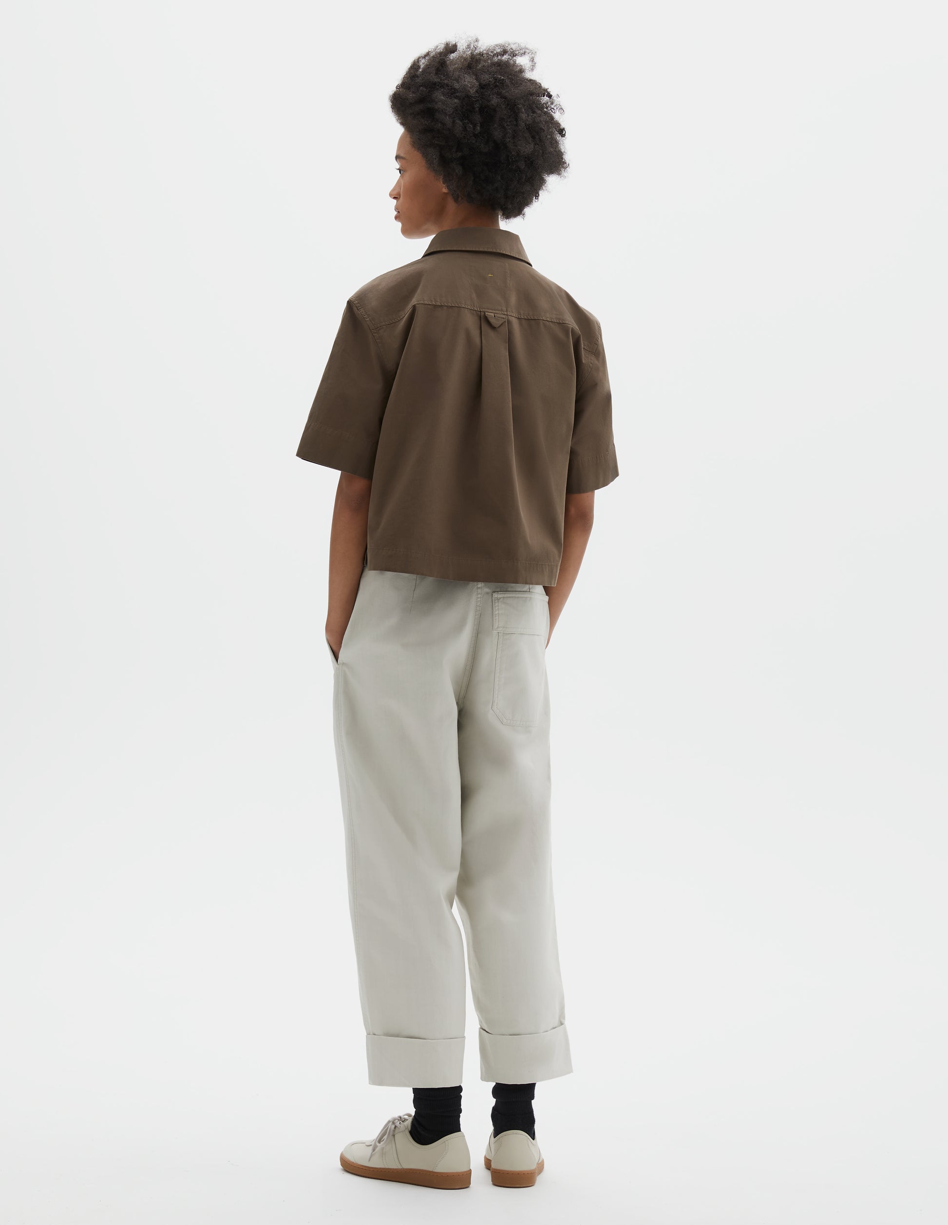 Full body image, back view of model wearing greenish brown cotton short sleeve shirt, with stepped side hem, hanger loop and box pleat on back yoke. Styled with white trousers, black socks and beige nubuck trainers.
