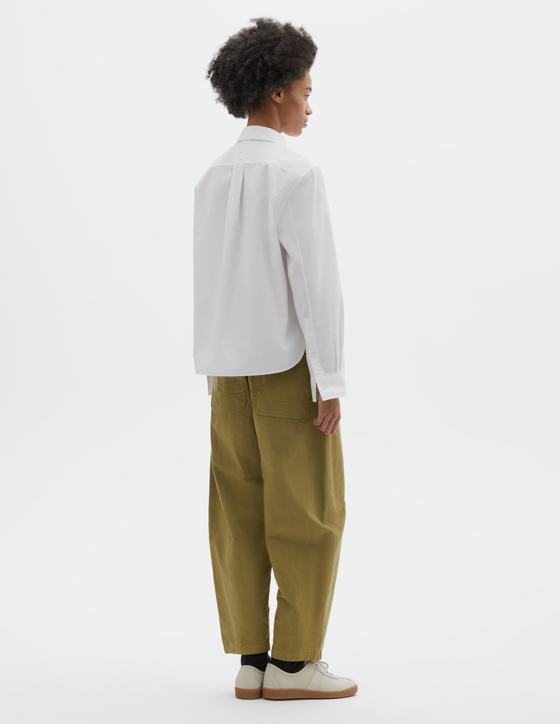 Full body image, back view of model wearing white regular cut shirt with box pleat on back yoke, corozo button cuffs and curved hem. Styled with green trousers, black socks and beige suede trainers.