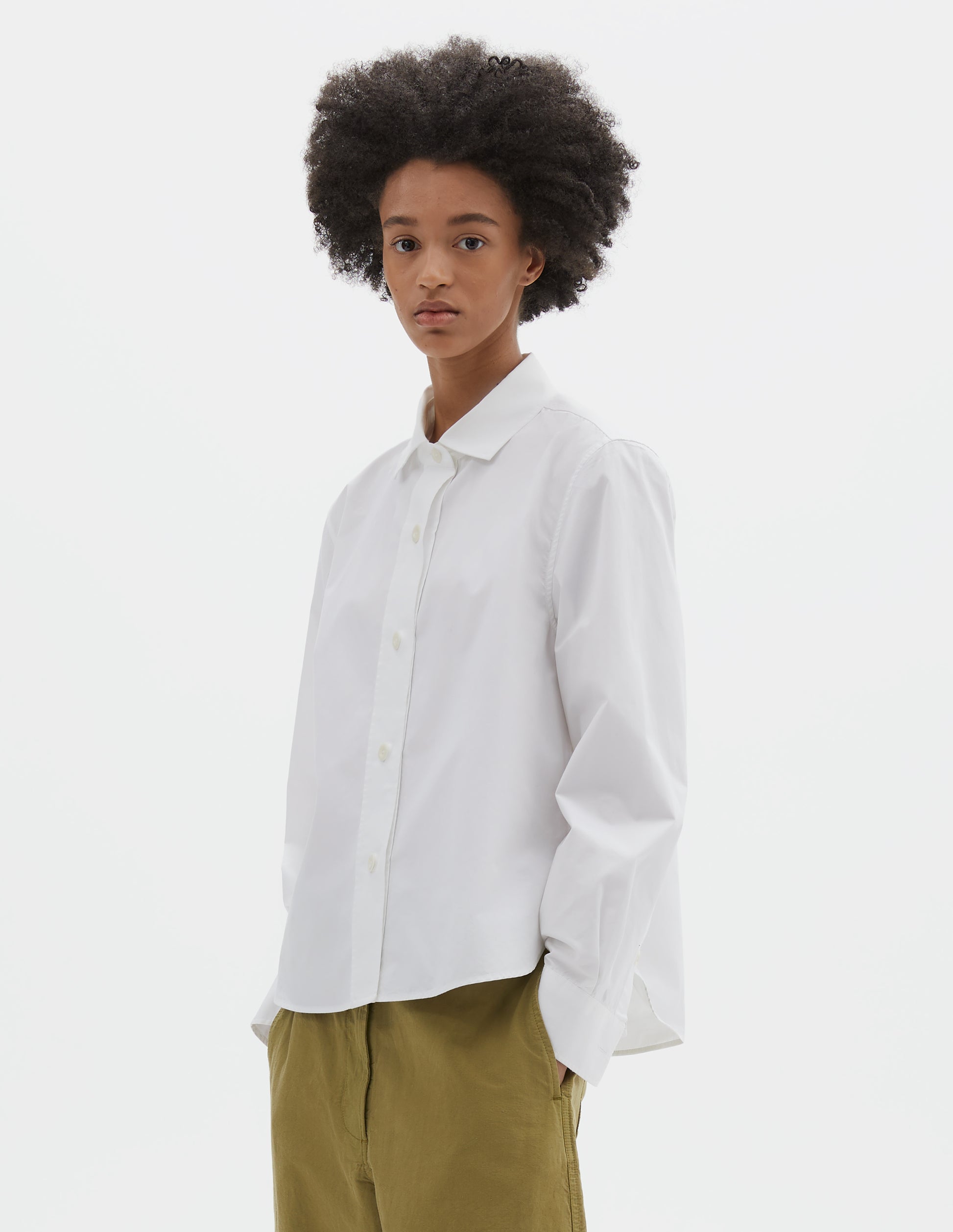 Cropped body image, front view of model wearing white regular cut shirt with stitch through raised placket, corozo buttons on placket and cuffs, curved hem. Styled with green trousers.