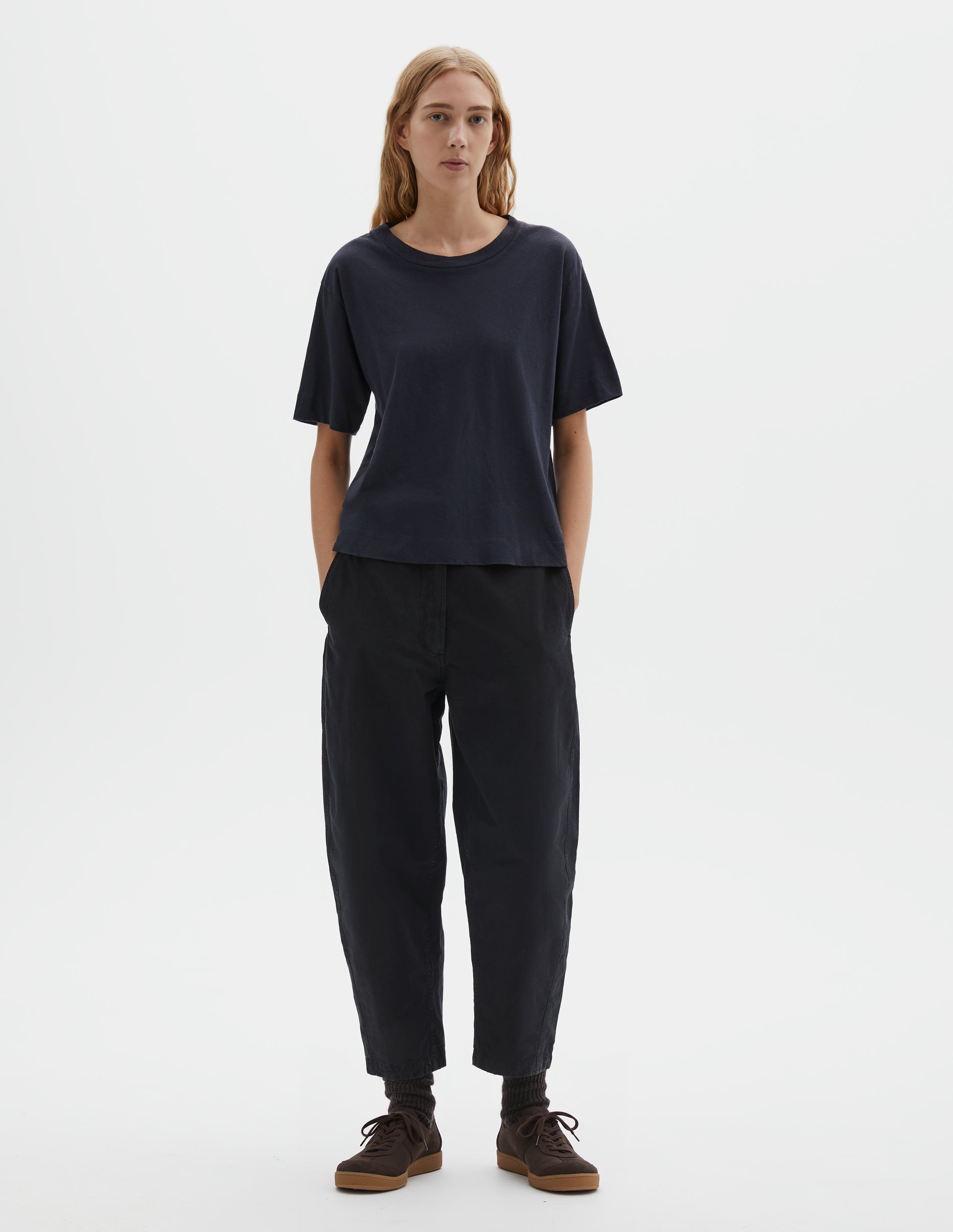 Full body image, front view of model wearing navy blue short sleeve cotton t-shirt with contrast rib on neck. Styled with indigo blue trousers, black socks and brown suede trainers.