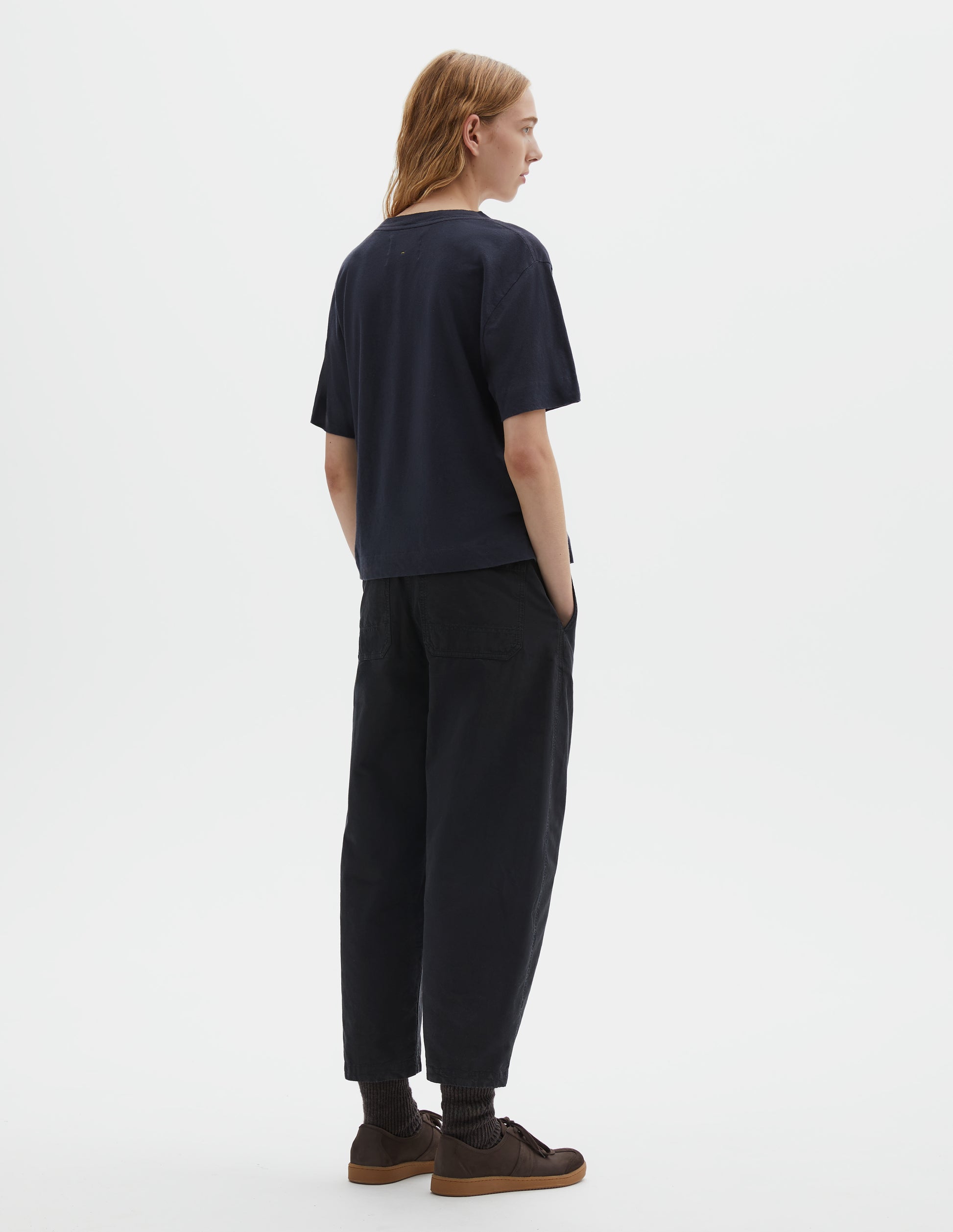 Full body image, back view of model wearing navy blue short sleeve cotton t-shirt with contrast rib on neck and stitch detailing on back shoulder. Styled with indigo blue trousers, black socks and brown suede trainers.
