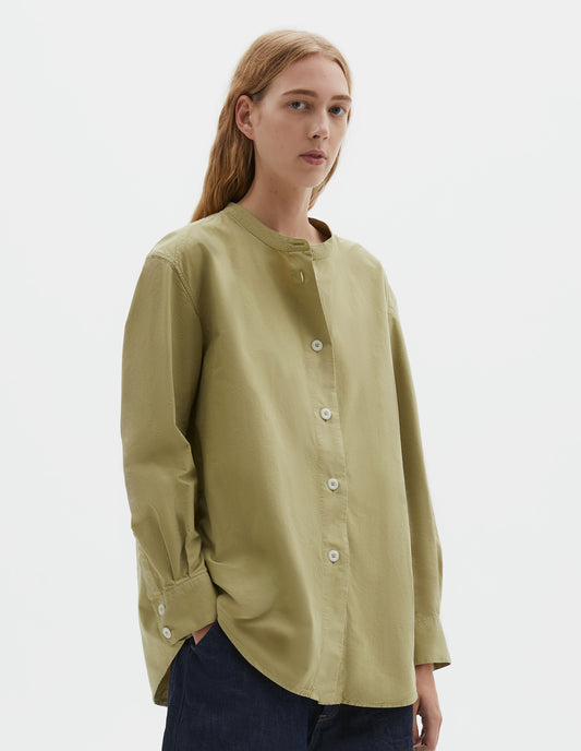 Cropped body image, front view of model wearing yellowish green cotton canvas long sleeve collarless shirt, with wide stitch through placket, corozo brace buttons and double button fastening cuffs. Styled with indigo blue denim shorts.