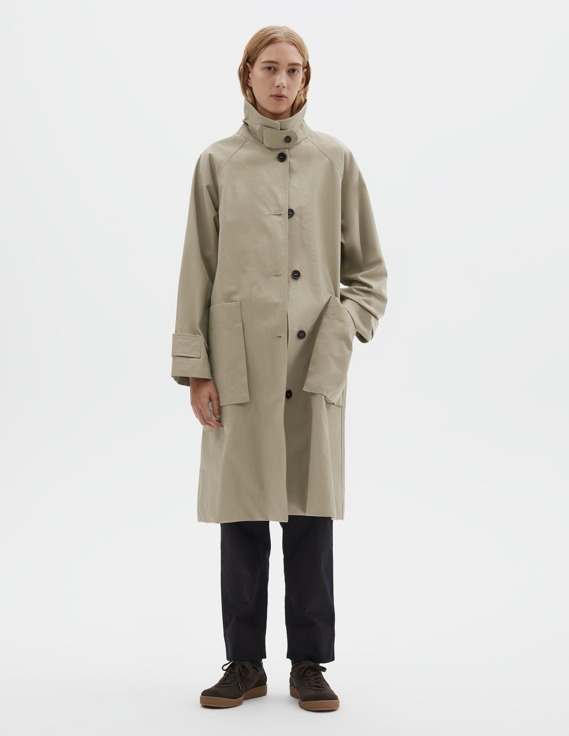 Full body image, front view of model wearing beige cotton canvas long sleeve mac, with storm tab fastening neck, raglan sleeve, button tab adjustable cuffs and large patch bellow pockets. Styled with collar popped, with navy blue trousers and brown suede trainers.