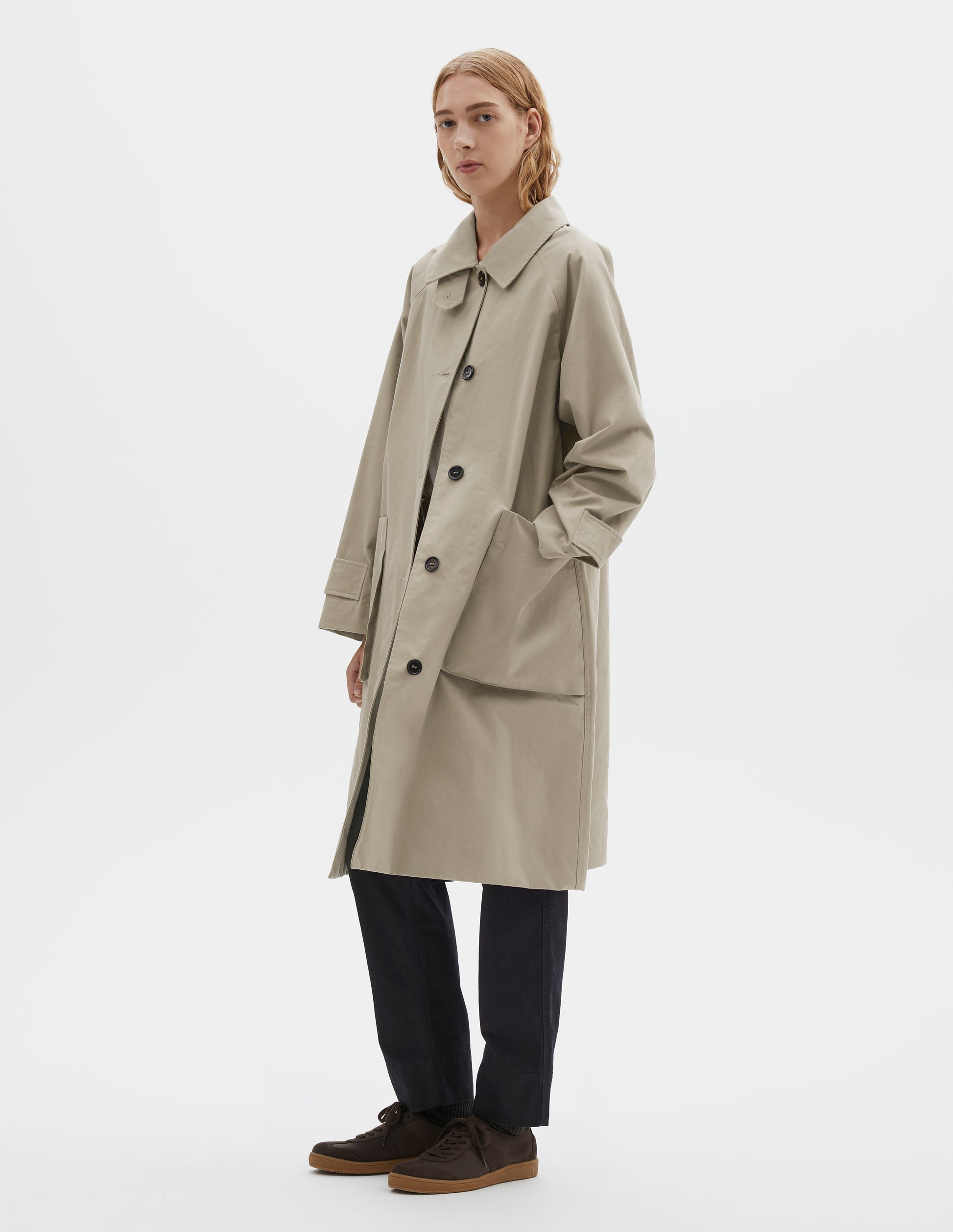 Full body image, side view of model wearing beige cotton canvas long sleeve mac, with storm tab fastening neck, raglan sleeve, button tab adjustable cuffs and large patch bellow pockets. Styled with navy blue trousers and brown suede trainers.