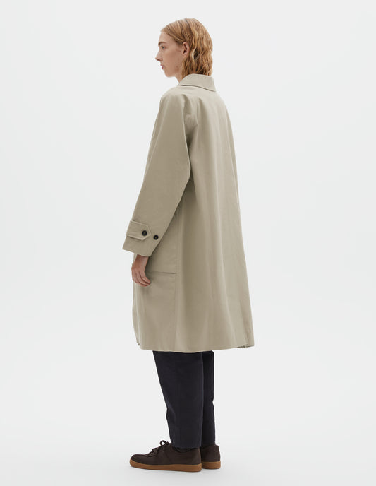 Full body image, back view of model wearing beige cotton canvas long sleeve mac, with button tab adjustable cuffs and rear vent. Styled with navy blue trousers and brown suede trainers.