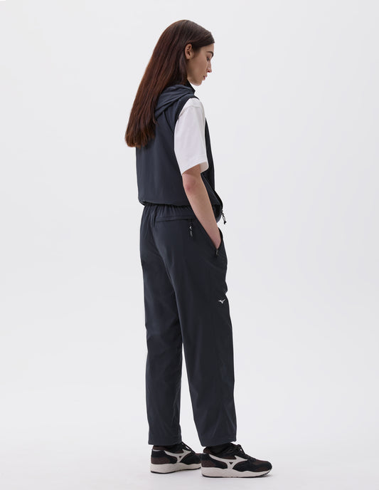 Full body image, back view of model wearing relaxed dark blue trousers with zip side pocket, zip welt back pocket and small white Mizuno logo, styled with dark blue gilet with hood, white t-shirt and black, brown and white trainers.