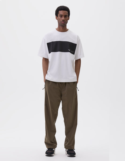 Full body image, front view of model wearing white t-shirt with black block running across chest, branded with Mizuno and Margaret Howell logo. Styled with green trousers with zipper pockets and black and brown trainers.