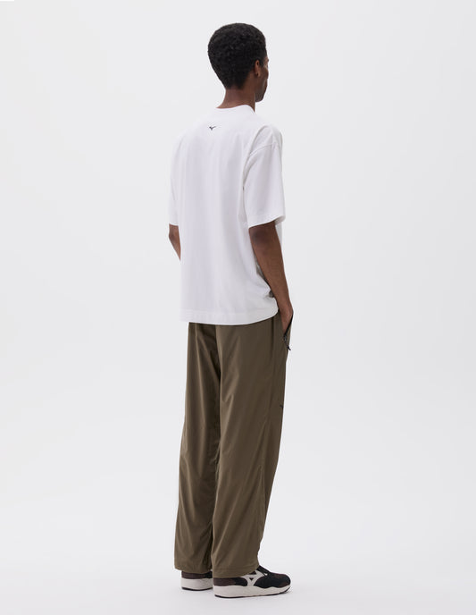 Full body image, back view of model wearing white t-shirt with black Mizuno logo on back of neck. Styled with green trousers with zipper pockets and black and brown trainers.