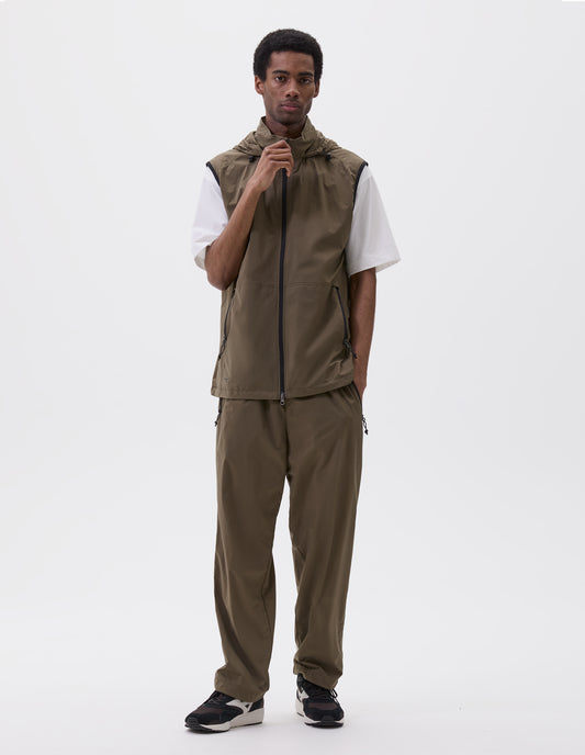 Full body image, front view of model wearing green/brown sleeveless gilet with adjustable hood, zip fastening, elasticated puller and concealed zip side pockets. Styled with white t-shirt, matching green/brown trousers and black and brown trainers.