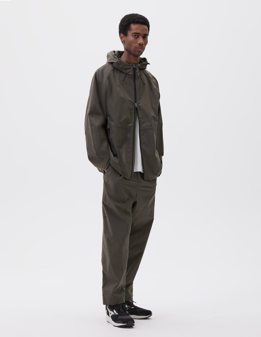 Full body image, front view of model wearing green/brown lightweight rain jacket with adjustable hood, zip fastening closure and pockets and elasticated hem. Styled with white t-shirt, matching green/brown trousers and brown and black trainers.