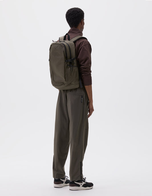 Full body image, back view of model wearing khaki green nylon ripstop backpack with side pockets, contrasting black carry handle and contrasting black chest belt. Styled with brown high neck top, green trousers and black and brown trainers.