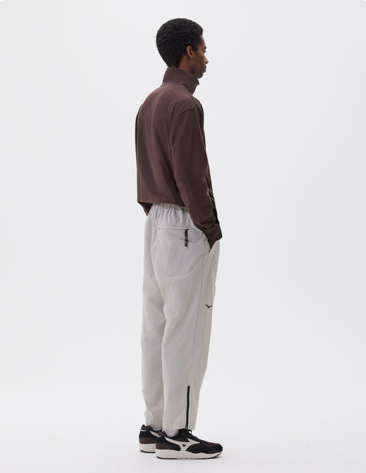 Full body image, back view of model wearing light grey lightweight trousers with elasticated waist with toggle cinch adjuster, zipper on lower leg and slant pockets. Styled with brown half zip sweatshirt and black and brown trainers.