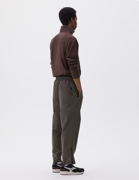 Full body image, back view of model wearing green/brown lightweight trousers with elasticated waist with toggle cinch adjuster, zipper on lower leg and slant pockets. Styled with brown half zip sweatshirt and black and brown trainers.