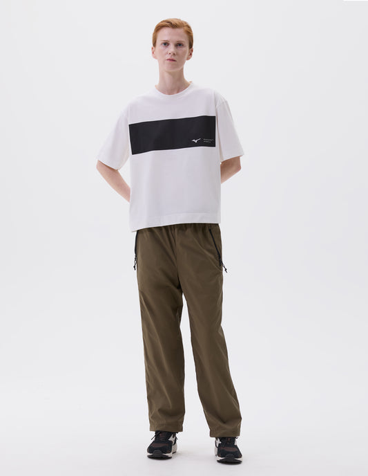 Full body image, front view of model wearing white relaxed t-shirt with white Mizuno and Margaret Howell logo on black wide stripe, styled with green trousers with zip side pockets and black, brown and white trainers.