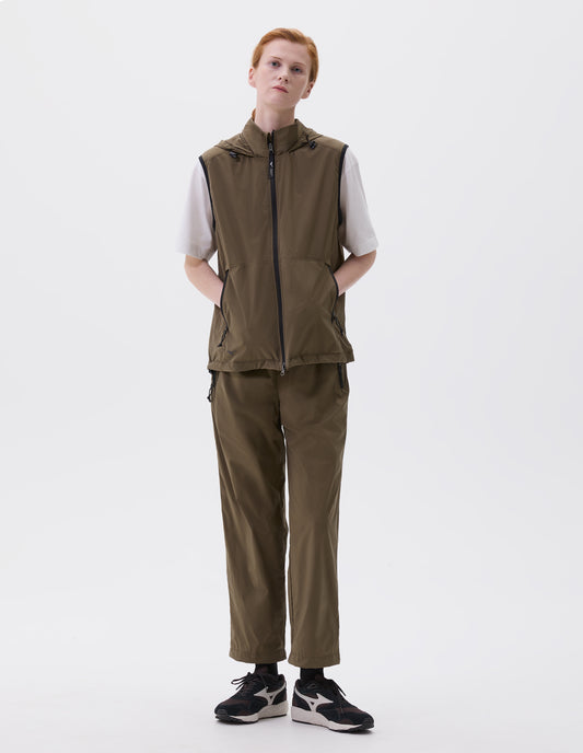 Full body image, front view of model wearing green regular cut gilet with zip side pockets, zip fastening, hood with elastic adjuster and small black Mizuno logo, styled with white t-shirt and green trousers, black socks and  with black, brown and white trainers.