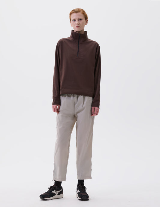 Full body image, front view of model wearing wearing brown relaxed cut high neck half zip sweatshirt, styled with light grey trousers and black socks with black, brown and white trainers.
