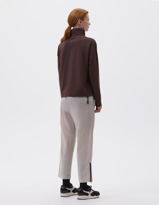 Full body image, back view of model wearing wearing brown relaxed cut high neck sweatshirt with small black Mizuno logo, styled with light grey trousers with zip on hem and black socks with black, brown and white trainers.