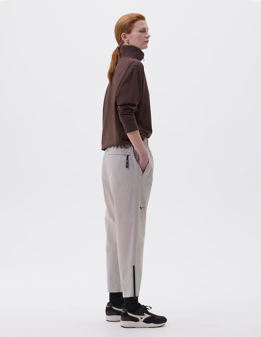 Full body image, back view of model wearing light grey relaxed cut trousers with side pockets and elasticated waist, zip welt back pocket, black zip on hem and small black Mizuno logo, styled with brown half zip sweatshirt, black socks with black, brown and white trainers.
